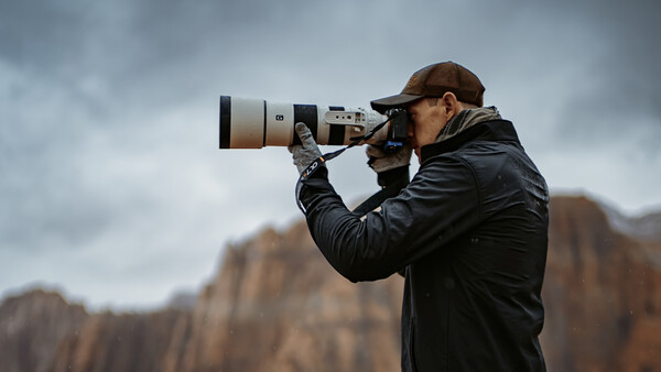 Photographer with telephoto lens pointed toward rocky mountain landscape under overcast sky.