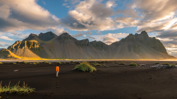 Solitary hiker in orange jacket on black sand beach beneath dramatic mountain peaks during golden hour.