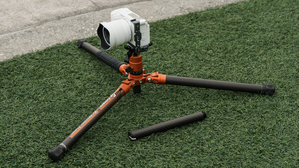 Orange and black camera tripod with white video camera mounted on green artificial turf backdrop.