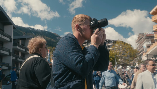 Photographer with camera raised to eye, focusing on subject during outdoor event with crowd and mountains in background.