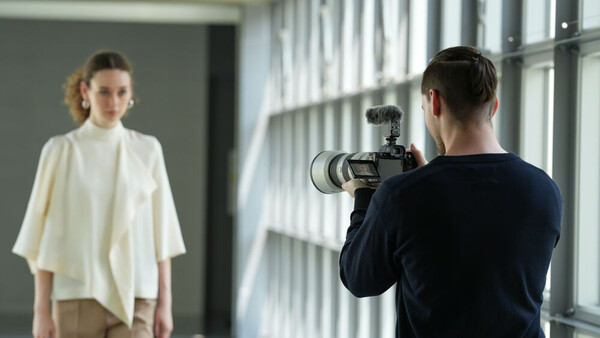 Photographer operating a camera on a shoulder rig while capturing a portrait of a woman in a bright, minimalist studio.