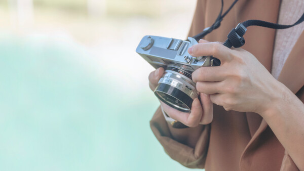 Hands holding a vintage rangefinder camera with neck strap against blurred turquoise background.