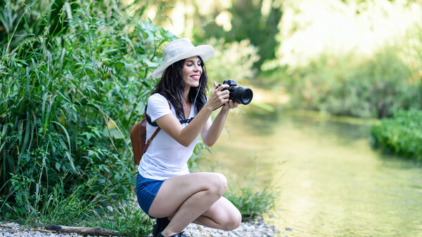 Woman crouching by a riverside holding a camera, wearing a white cap and casual clothing.
