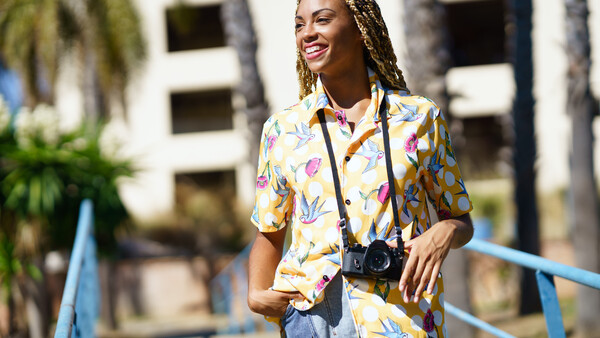 Smiling woman wearing a colorful floral shirt and camera on a lanyard, standing outdoors near palm trees.