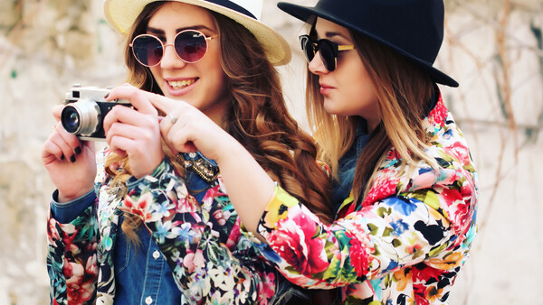 Two women in colorful floral blazers and hats holding a vintage camera between them.
