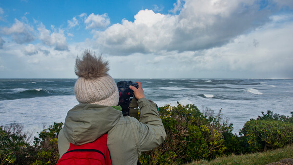 Person in winter hat and jacket photographing ocean waves from a coastal clifftop.