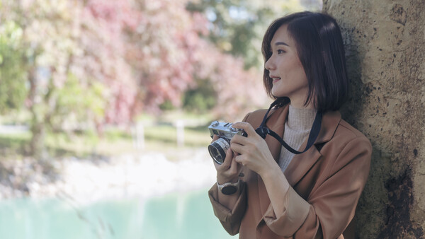 Woman holding a vintage rangefinder camera while leaning against a tree with blooming flowers in soft focus background.
