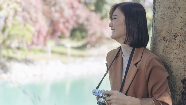 Woman holding a vintage camera, leaning against a textured wall with blooming flowers in soft focus behind her.