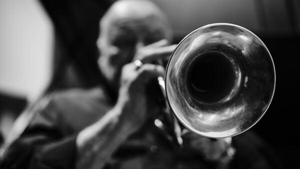 Musician playing trumpet, photographed from front with instrument bell in sharp focus and player blurred in background.