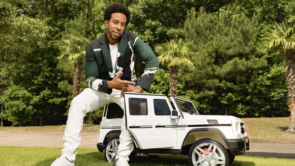 Man in green jacket and white pants posing with a miniature white SUV on grass with trees in background.