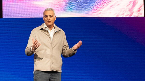 Man presenting on stage with hands gesturing while speaking before a blue backdrop with colorful digital art.