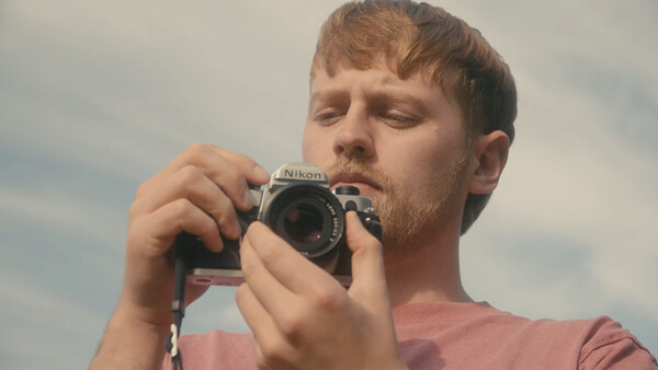 Male photographer holding a Nikon camera to his face while looking downward against a cloudy sky.