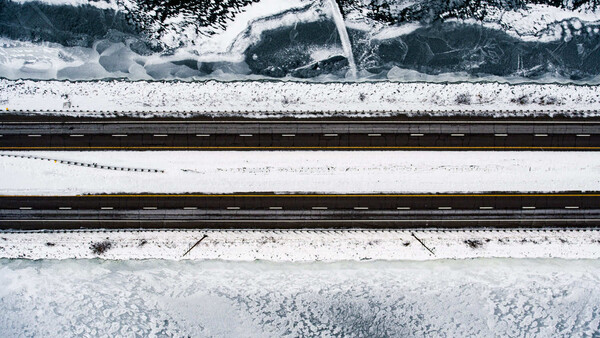 Aerial view of snow-covered landscape with railroad tracks and forested areas separated by white fields.