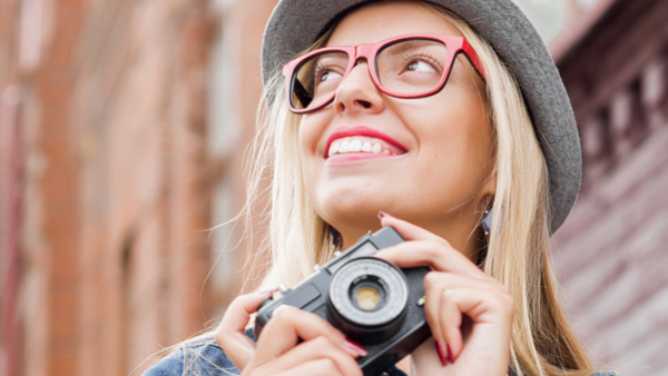 Young woman wearing pink-framed glasses and gray beanie holding vintage camera, smiling upward against brick wall backdrop.