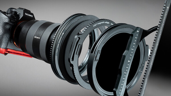 Sony mirrorless camera with multiple lens filters and adapters arranged in front of it against a gray background.