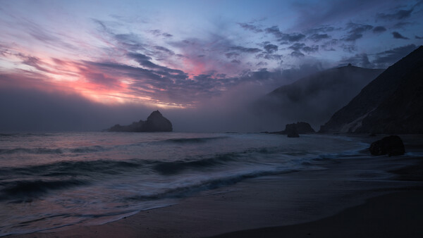 Dramatic sunset over a rocky coastline with dark silhouetted cliffs framing calm ocean waves.