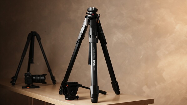 Professional tripod with ball head and video camera on wooden surface against warm backdrop.