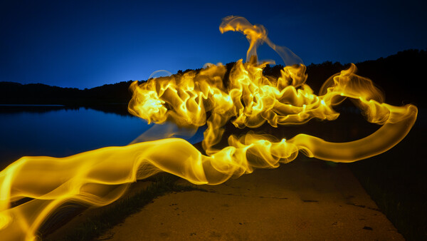 Golden silk fabric flowing across a dark beach at twilight with calm water and mountains in the background.