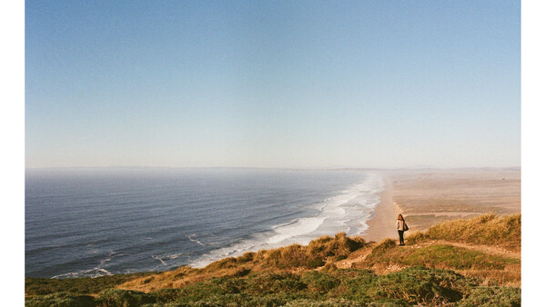 Hiker standing on golden clifftop overlooking a curved coastline with white-capped waves and blue ocean.