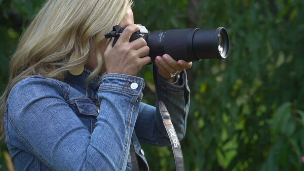 Photographer with blonde hair using a telephoto lens, wearing a denim jacket and sunglasses.