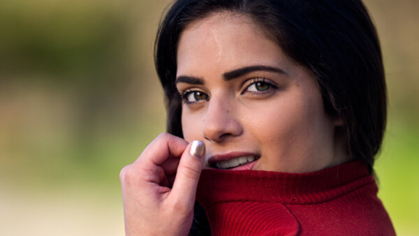 Woman with dark hair wearing a burgundy turtleneck, hand near face, smiling at camera with soft natural lighting.