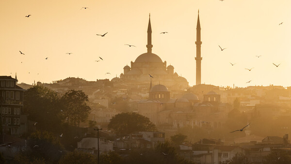 Mosque with dual minarets silhouetted against golden sunset sky with birds in flight.