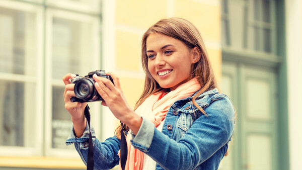 Smiling woman holding a DSLR camera with a telephoto lens, wearing a denim jacket and coral scarf.