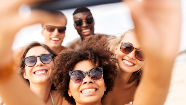 Group of five smiling people wearing sunglasses taking a selfie outdoors in bright daylight.