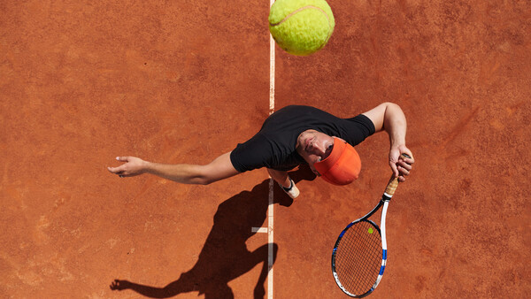 Tennis player executing an overhead serve on a clay court, captured from above with the ball suspended above the net line.