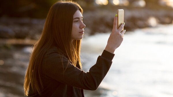 Woman holding smartphone horizontally while standing outdoors near water during golden hour.