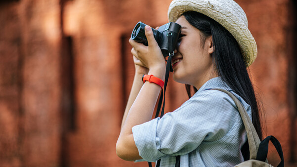 Female photographer wearing a straw hat and light blue shirt holding a camera to her face while photographing against a blurred brick wall background.