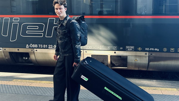 Person in dark formal attire standing beside a black coffin on a train platform next to a passenger train.