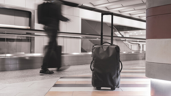 Black rolling luggage sits stationary on a tiled floor while a blurred figure walks past in a modern airport terminal.