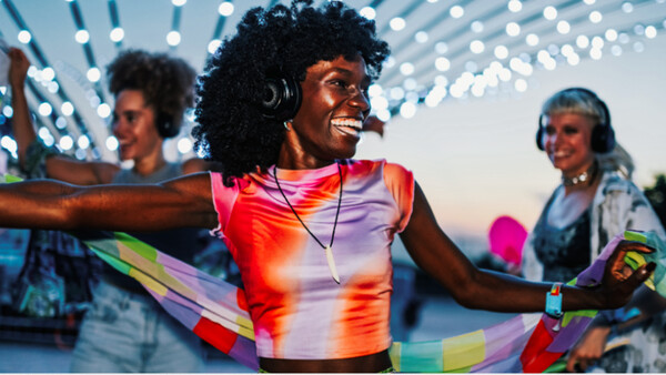 Woman wearing colorful gradient tank top and headphones dances outdoors at twilight under string lights.