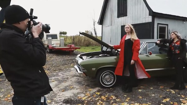Photographer shooting a woman in a red coat posing by a vintage car with open hood.