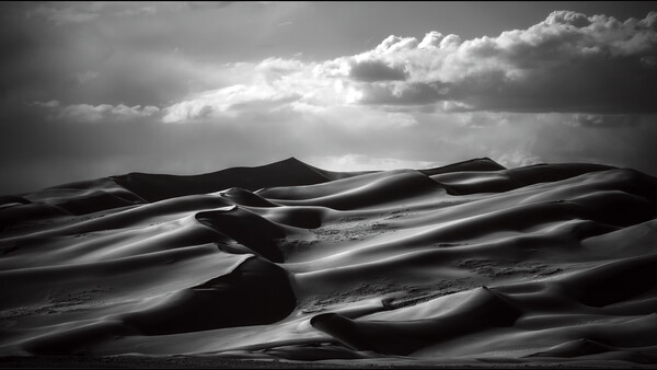 Monochromatic desert landscape with undulating sand dunes beneath dramatic clouded sky with light rays.