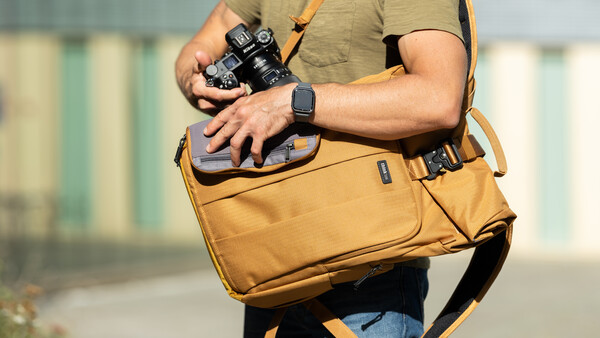Photographer holding a digital camera while wearing a tan messenger bag and smartwatch.