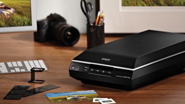 Flatbed scanner on wooden desk with photo prints, camera, and studio setup visible in background.