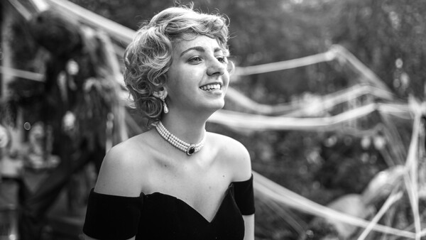 Woman in off-shoulder black dress with pearl necklace, looking upward with a smile in a black and white photograph.