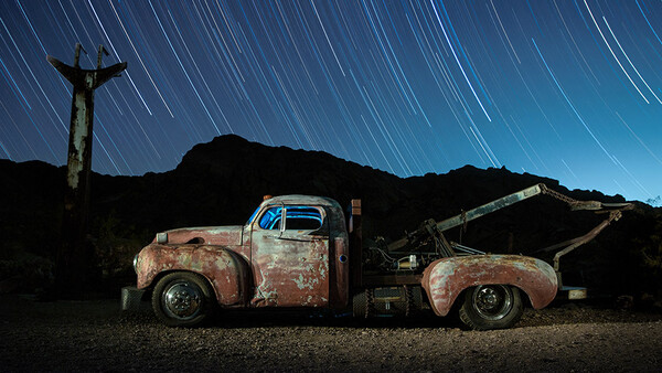 Two rusted vintage pickup trucks positioned side-by-side in a desert landscape at night, with star trails streaking across the dark sky and a cross silhouetted on a distant hill.