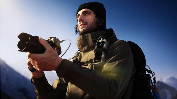 Male photographer holding a camera against a clear blue sky with mountains in the background.