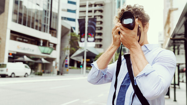 Man in light blue shirt holding camera to his face on an urban street.