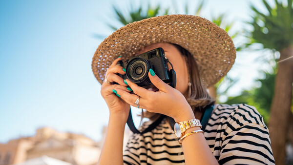 Woman in straw hat and striped shirt holding film camera up to her face.