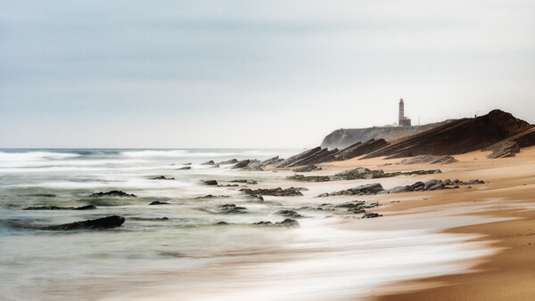 Long exposure seascape with blurred waves, rocky shore, and distant lighthouse on coastal headland.