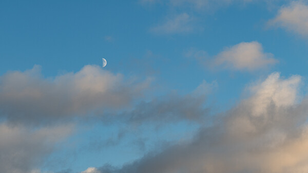 Crescent moon visible in daylight sky among white and gray clouds.