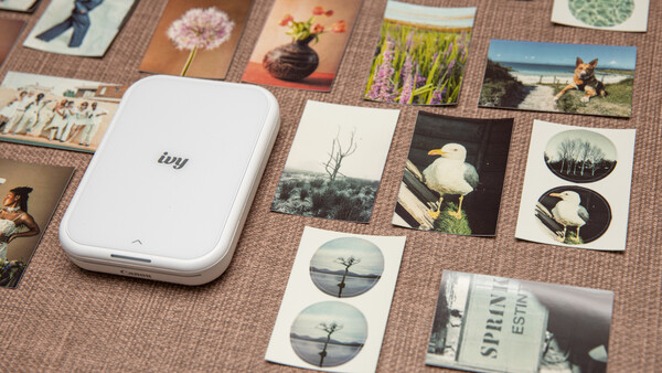 Portable photo printer surrounded by printed photos and contact sheets on a textured surface.