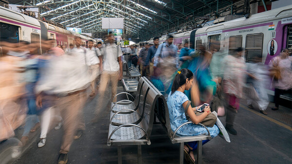 Woman sitting on a bench at a train station, surrounded by blurred commuters and purple trains.