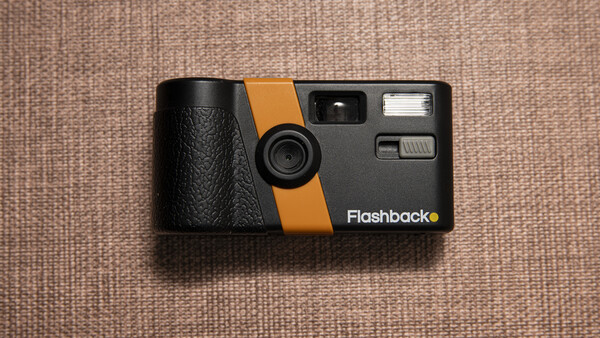 Flashback vintage film camera with black body and orange stripe on beige fabric background.