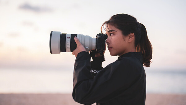 Photographer with long dark hair using a telephoto lens during golden hour at a coastal location.