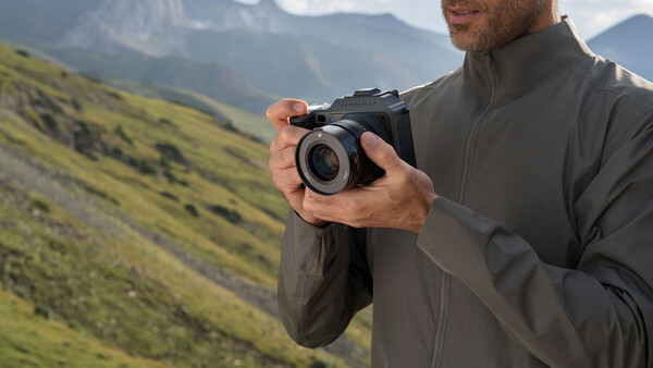 Photographer holding a mirrorless camera while standing in a mountainous landscape with green slopes.